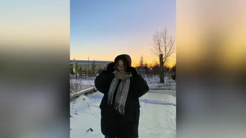 Woman in a black winter coat and plaid scarf standing in a snowy landscape during a golden sunset.