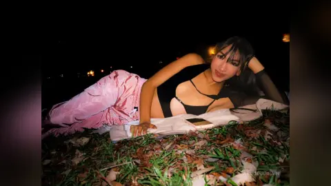 Young woman in a black crop top and pink satin trousers relaxing on grass at night.