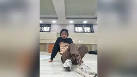 Young woman in casual black sweatshirt and brown trousers sitting relaxed on a floor in a modern room.