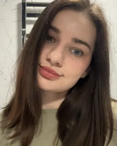 Close-up portrait of a young woman with long brown hair and a natural makeup look in soft indoor lighting.