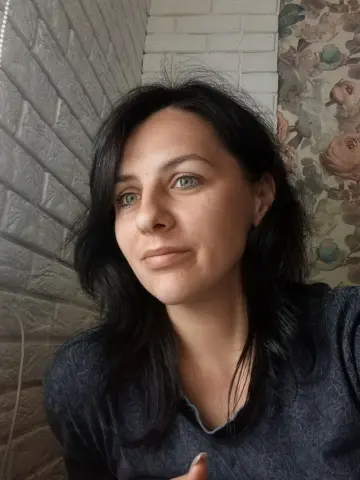 Portrait of a woman with dark hair and green eyes wearing a grey shirt in a room with brick walls.