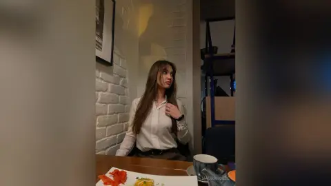 Young woman in a white blouse sitting at a cafe table with a thoughtful expression near a white brick wall.