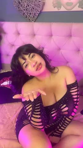 Young woman in black cutout bodysuit relaxing on a pink tufted headboard in soft purple lighting.