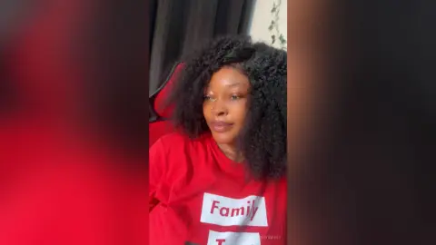 Smiling young woman with curly hair wearing a red graphic t-shirt in a cozy indoor setting.