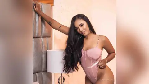 Smiling woman in a pink lace bodysuit posing in a cozy bedroom setting.
