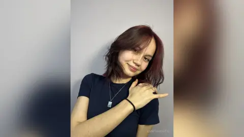 Smiling young woman with wavy brown hair wearing a navy blue t-shirt against a plain grey studio background.