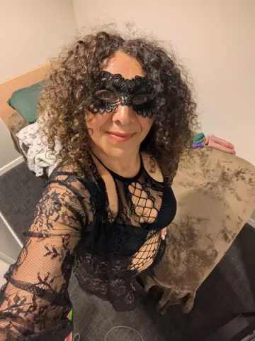 Smiling woman with curly hair wearing a black lace masquerade mask and sheer lace outfit in a cozy bedroom.