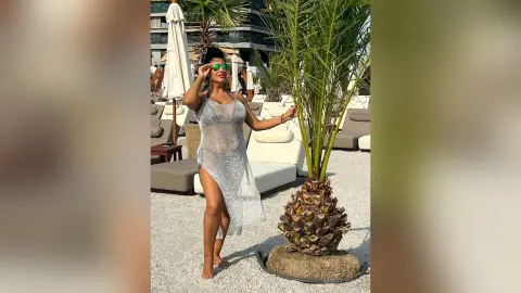 MissyJolie stands outdoors in a sheer, shimmering silver mesh dress, wearing green sunglasses and posing next to a small palm tree.