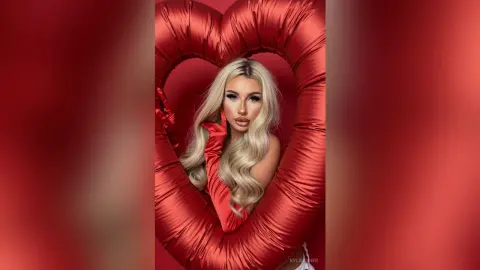 Glamorous blonde woman in red satin gloves posing inside a large red heart prop for a romantic photoshoot.