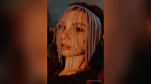Woman with red hair wearing a silver chain headpiece in a moody, cinematic portrait.