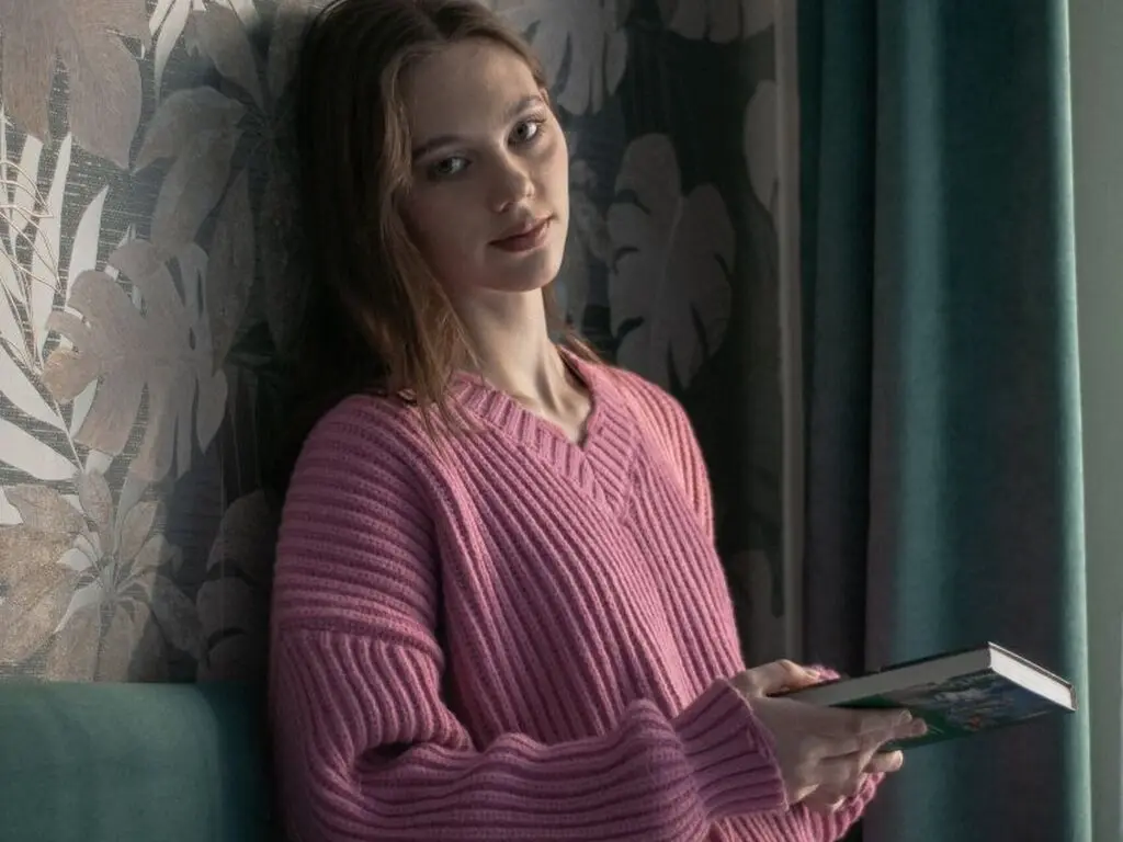 Young woman in a pink ribbed sweater reading a book against floral wallpaper in a cozy, moody room.