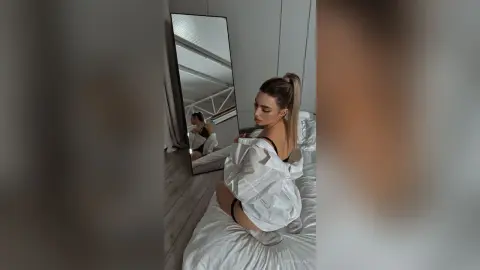 Young woman in a white oversized shirt posing in a minimalist bedroom with a large floor mirror.