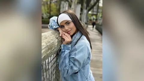 Smiling young woman in a denim jacket and white headband leaning on a wooden fence in a sunny outdoor park.