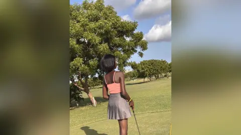 Young woman in a peach tank top and grey skirt standing in a sunny green field by a large tree.