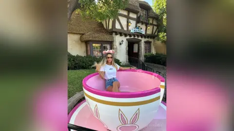 MiaGuerra wears a white t-shirt, denim shorts, and pink bunny ears while sitting inside a large pink and white teacup outdoors.