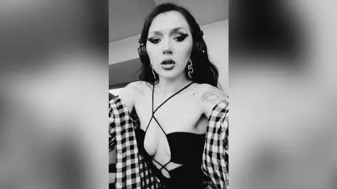 Young woman with winged eyeliner in a black strappy top and plaid shirt wearing headphones in a moody black and white portrait.