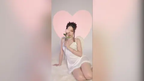 Dreamy woman in a white angel dress holding a pink rose against a soft heart background.