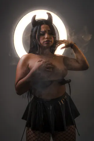 Woman in devil horns and black skirt posing with smoke in a moody, cinematic studio setting.