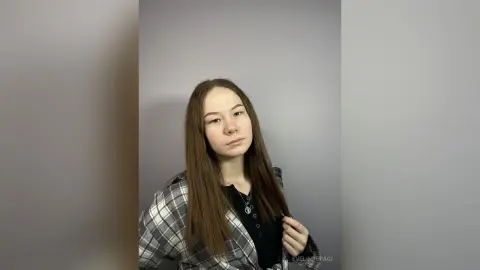 Young woman with long brown hair wearing a black top and grey plaid shirt against a neutral grey background.