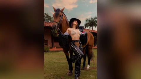 Stylish woman in black western outfit and hat posing with a brown horse in a rustic outdoor ranch setting.