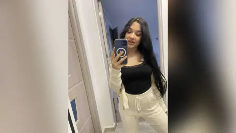 Young woman with long dark hair in a black bodysuit and cream lounge pants taking a mirror selfie at home.