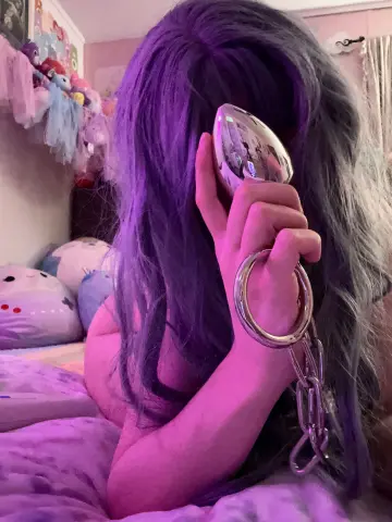 Woman with long purple hair wearing silver hoop earrings in a cozy, pink-toned bedroom setting.