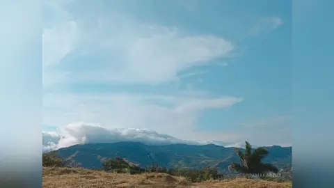 SamaraBert overlooking a vast, rolling mountain range under a bright blue sky with wispy white clouds.