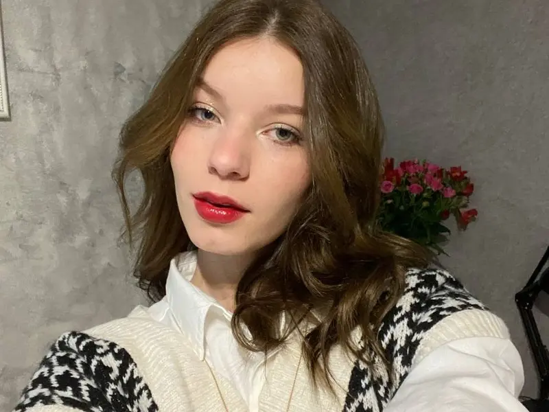 MYasmina poses with long brown hair and red lipstick, wearing a black satin top against a grey background.