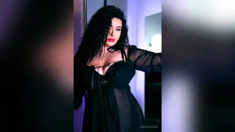 AdiraMaeven poses in a sheer black mesh outfit that shows her cleavage and skin, with her long dark wavy hair falling over her shoulders.