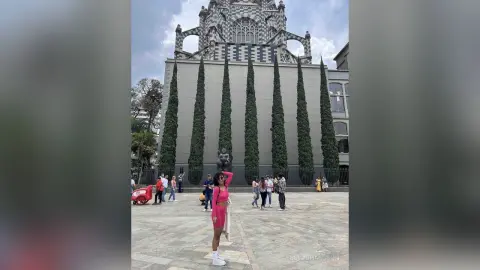 MissyJohansson stands in a plaza wearing a tight pink bodysuit and white sneakers, posing with one hand behind her head in front of tall, thin trees and a large building.