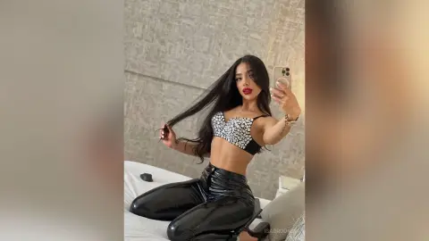 Young woman in a black sequin bralette and shiny leather pants posing confidently in a modern bedroom.