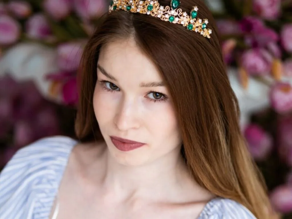 ZoeCao wears a gold and green tiara in her long brown hair while posing in front of pink flowers.