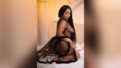 Glamorous woman in a sheer black mesh dress posing on a white bed in a luxury bedroom setting.