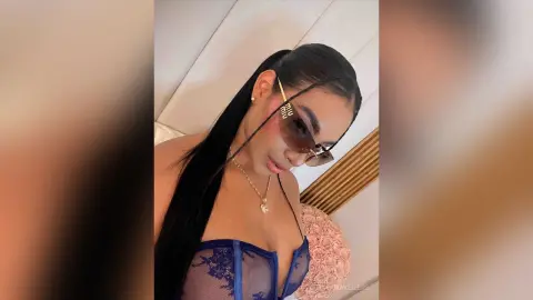Stylish woman with long dark hair wearing sunglasses and a blue lace corset in a bright indoor setting.