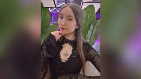 Young woman in a black lace long-sleeve top posing gracefully amidst lush green tropical leaves.