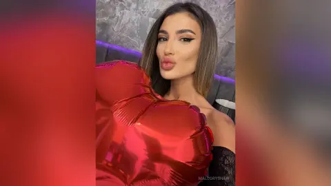 Glamorous woman with winged eyeliner posing with a large red heart balloon in a moody setting.