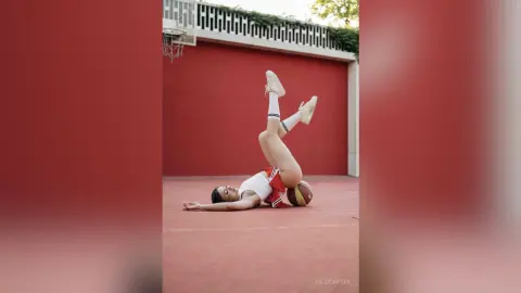 Athletic woman in white tank top and red shorts lying on a red basketball court with legs raised in a playful mood.
