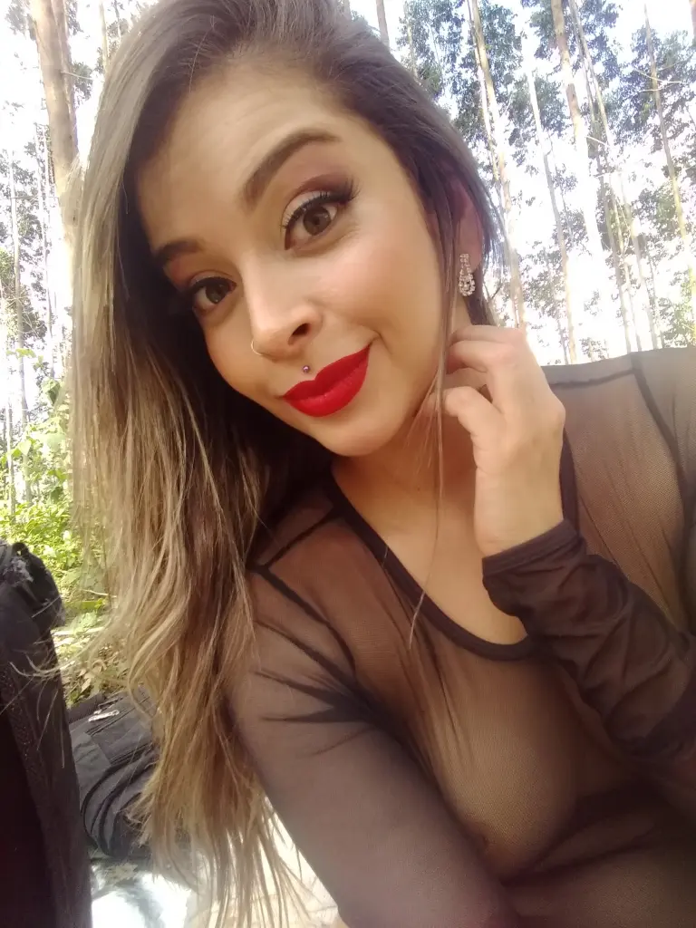 ChloeMackenziee with long brown hair and red lipstick, looking directly at the camera in a bright outdoor setting.