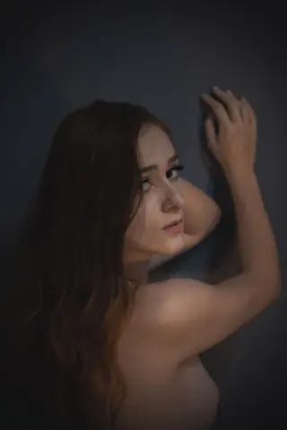 Moody portrait of a long-haired woman posing against a dark wall in soft, cinematic lighting.