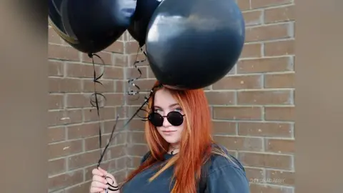 Redheaded woman in sunglasses holding black balloons against a brick wall for a moody aesthetic.