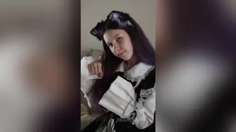 Young woman with purple hair wearing a black and white gothic lolita dress in a soft indoor setting.