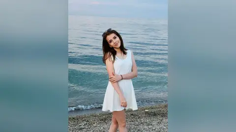Smiling young woman in a white summer dress standing on a pebble beach by the ocean under a clear sky.