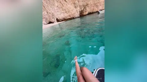 Clear turquoise ocean water near a rocky cliffside beach under bright sunlight.