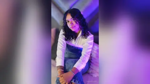 NaomiVelour poses wearing a white long-sleeve shirt and denim overalls, sitting with her dark curly hair down.