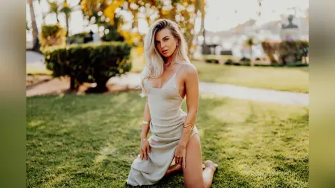 Blonde woman in a white summer dress kneeling on a sunny green lawn in a peaceful garden setting.