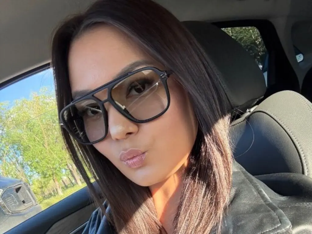 Stylish woman in black sunglasses and leather jacket sitting in a car on a sunny day.