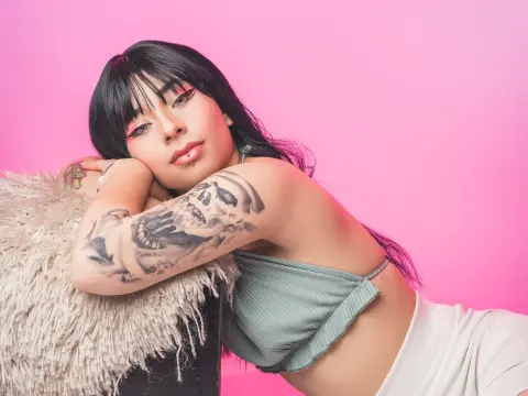 Young woman with black bangs and tattoos relaxing on a fuzzy pillow against a pink background.