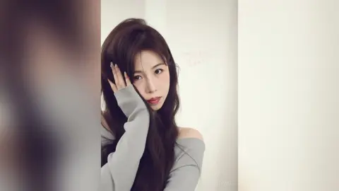 Young woman with long dark hair wearing a grey off-the-shoulder sweater in a soft, minimalist indoor setting.