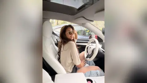 Smiling woman in a beige sweater sitting in a white Tesla car interior with sunlight filtering through the sunroof.