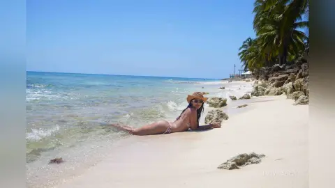 LucyMartin lies on her stomach on a white sand beach wearing a bikini and a straw hat.